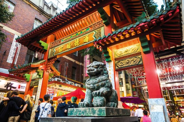 A weathered stone lion sits beneath a vibrant red and gold Chinese archway; people stroll through a bustling Chinatown street