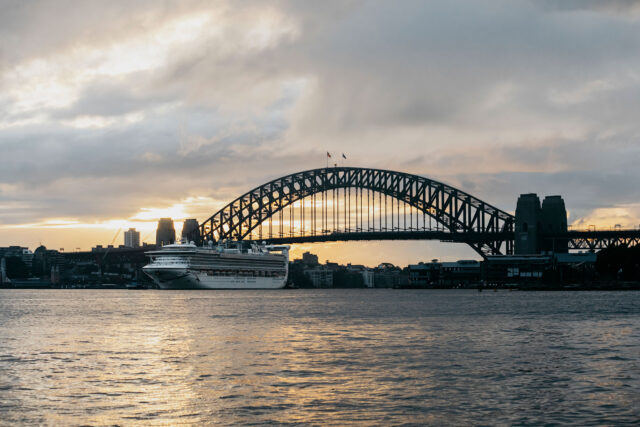 A cruise ship is docked under a large steel arch bridge during sunset