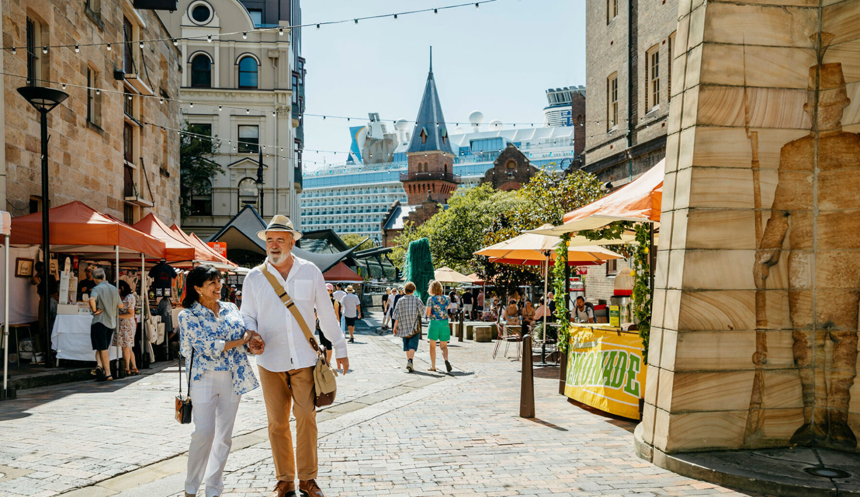 An elderly couple strolls hand-in-hand through a sunny cobblestone market square, past market stalls and outdoor seating