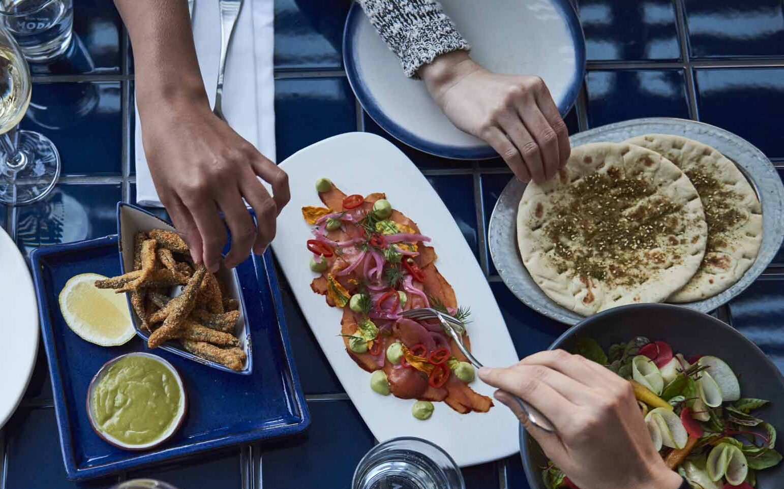 Hands reach for various dishes: fried fish, salmon with avocado, flatbreads, and a salad