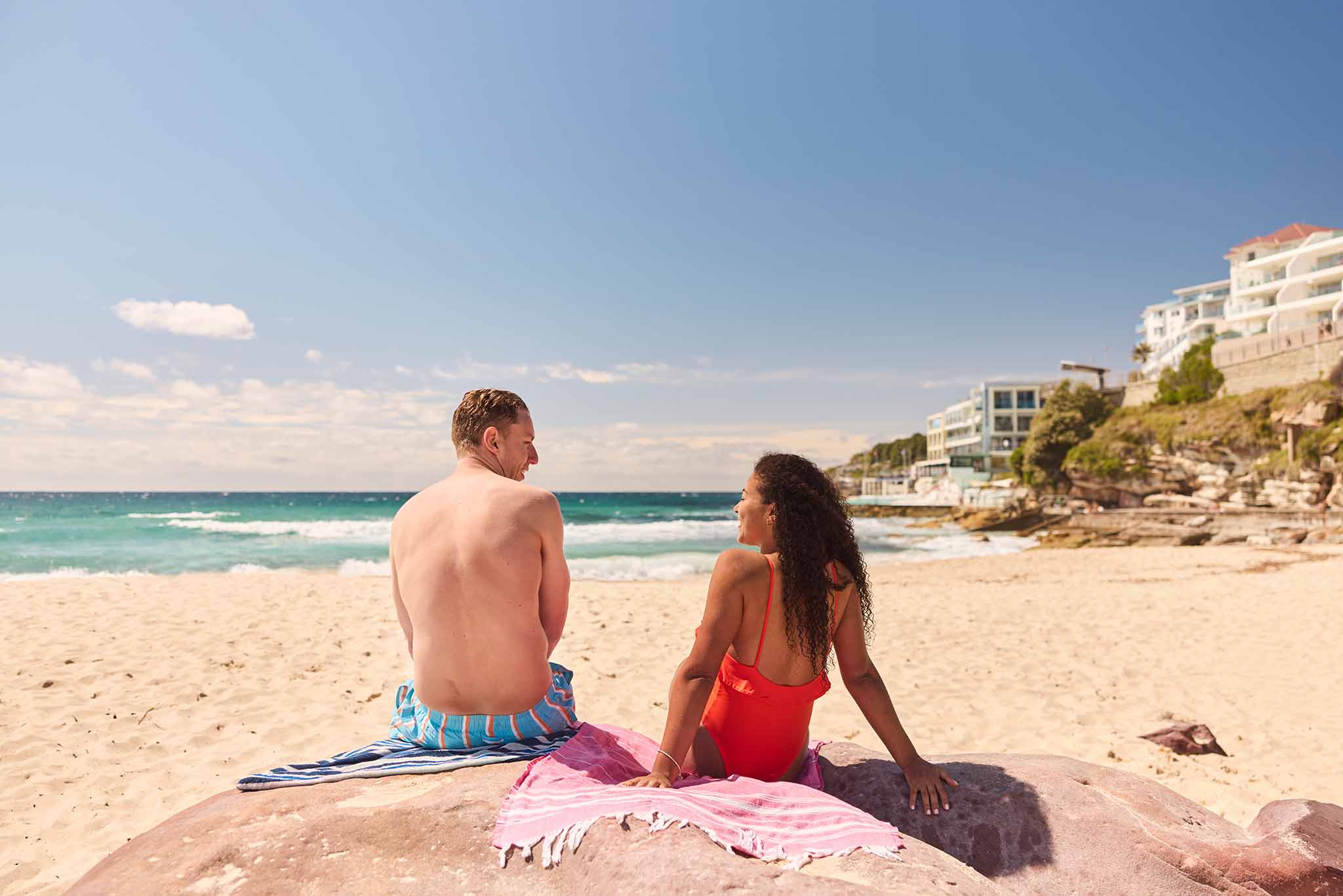 A couple sits on a rock, facing each other, on a sunny beach near buildings