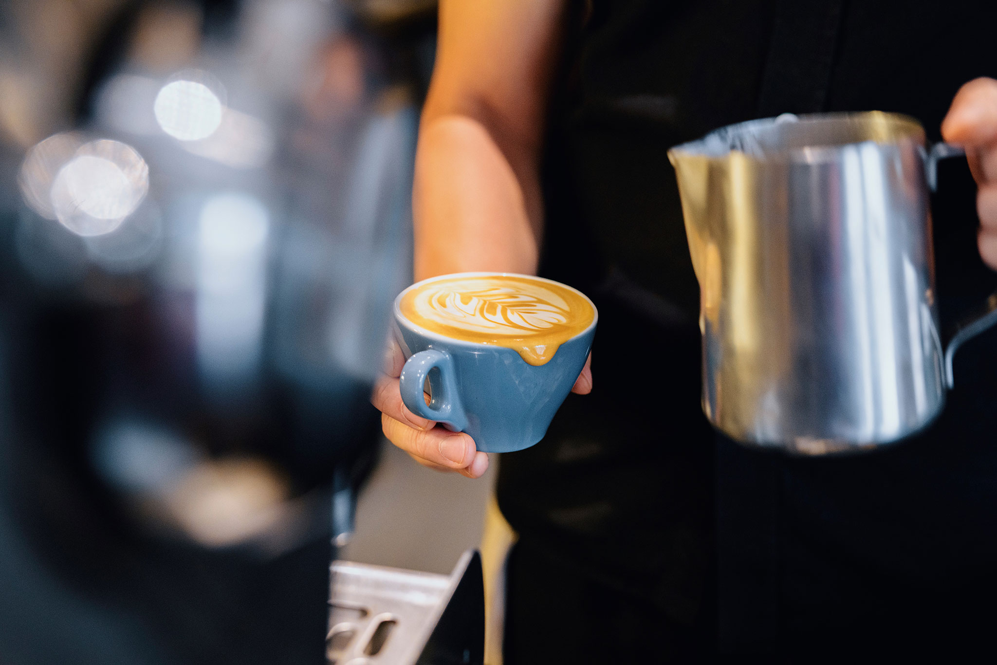 A barista holds a blue cup of latte art; they are pouring from a metal pitcher; the setting appears to be a coffee shop