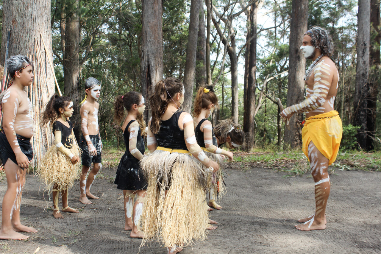 Children and a man, adorned with body paint and traditional attire, participate in a ceremony or lesson in a wooded area