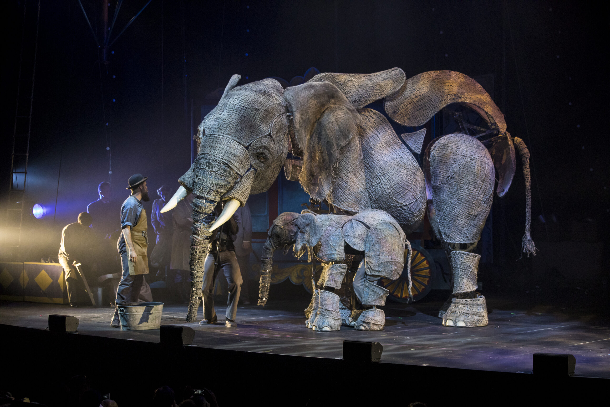 Large burlap elephant puppets are being manipulated by puppeteers on a dark stage
