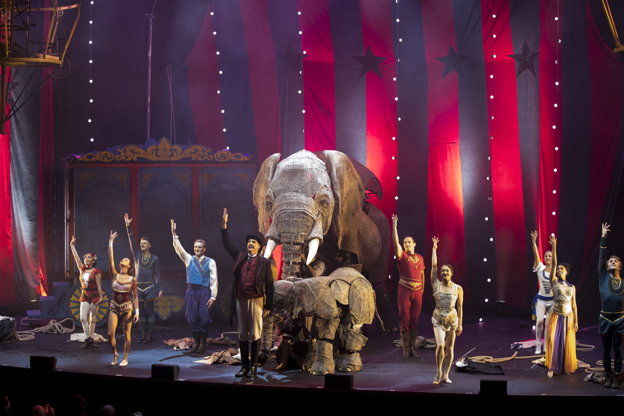 A large elephant puppet, flanked by performers, is presented on a circus stage
