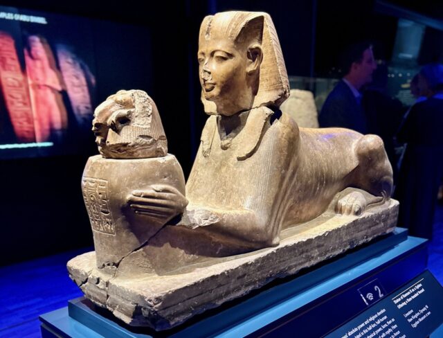 A sphinx statue, reclining, holding a smaller container; displayed in a museum setting, on a dark blue pedestal; the label reads: “2