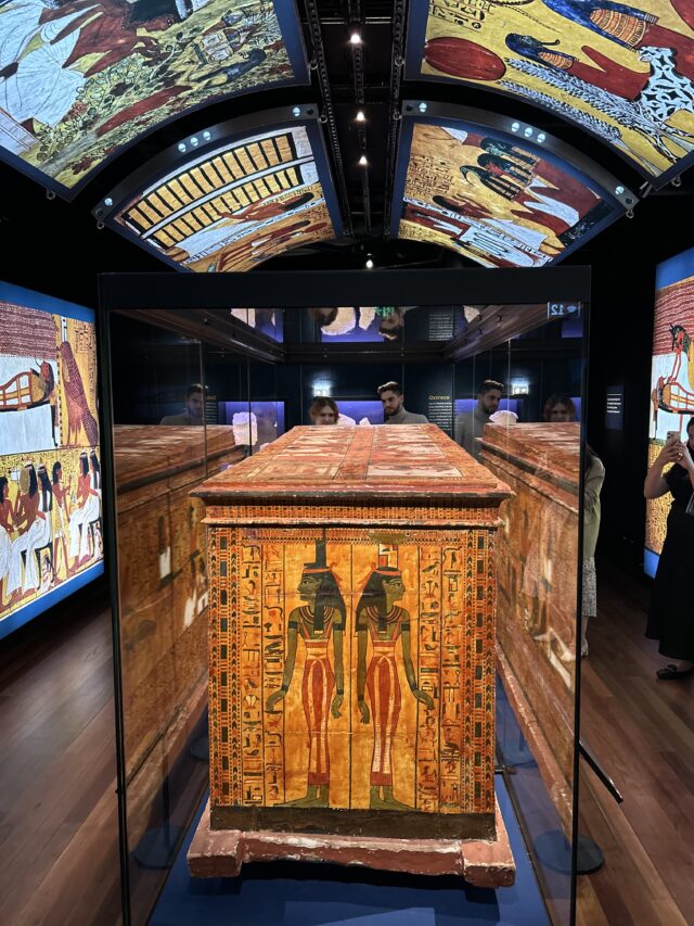 An ornately decorated ancient Egyptian sarcophagus, displaying painted figures, sits in a glass case