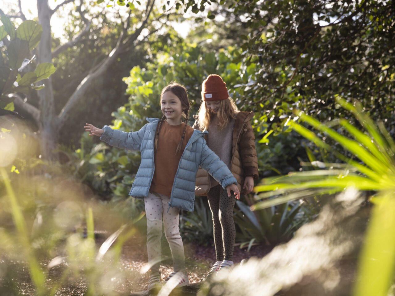 Two girls, wearing winter jackets, playfully balance on a log in a lush garden