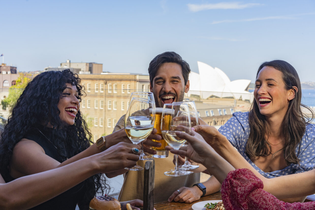 Four friends joyfully clink wine and beer glasses on a rooftop patio overlooking a cityscape and the Sydney Opera House