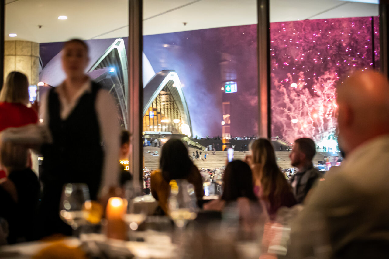 A server attends diners at a restaurant; a fireworks display and the Sydney Opera House are visible through a large window
