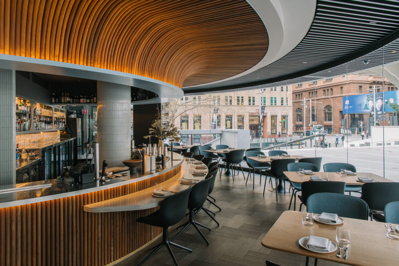 A curved bar with a wooden facade serves patrons seated at tables in a modern restaurant