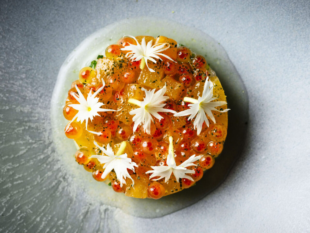 A salmon dish, adorned with white flowers and orange roe, rests on a gray plate