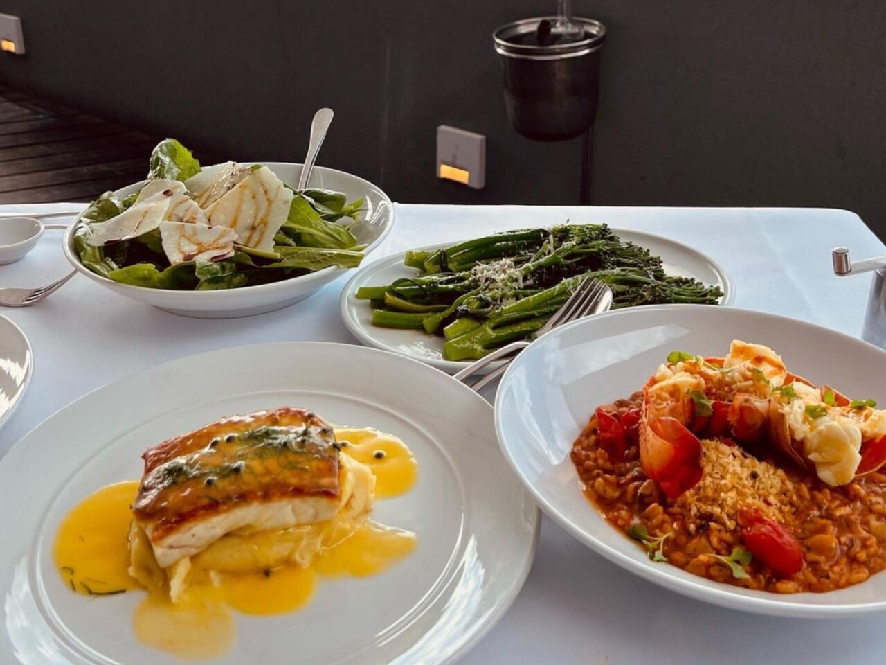 Pan-seared fish rests atop mashed potatoes and a yellow sauce; broccolini, salad, and lobster risotto are nearby on a white tablecloth