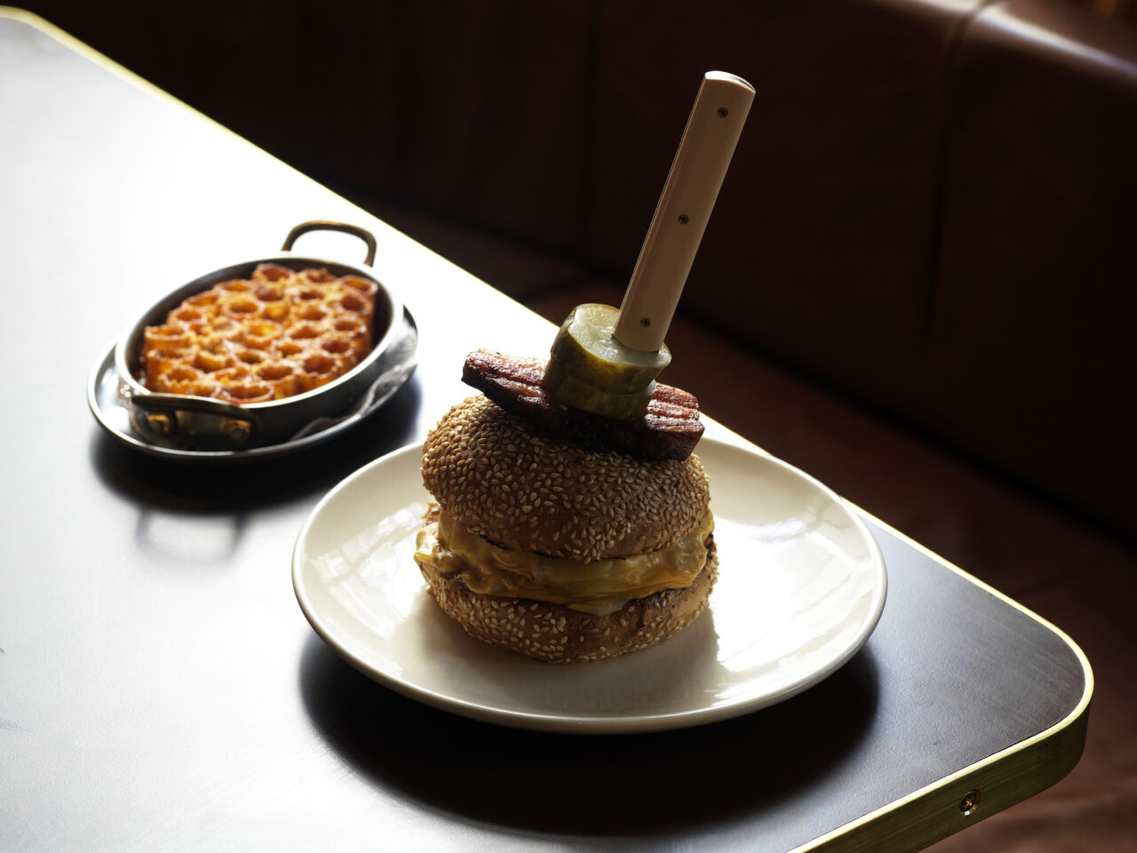 A hamburger, topped with bacon, a pickle, and a wooden skewer, sits on a plate