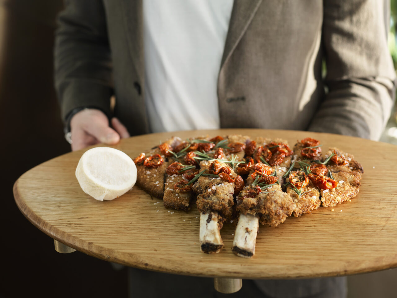 A breaded, bone-in meat dish, garnished with roasted tomatoes and rosemary, is presented on a wooden board by a person wearing a blazer