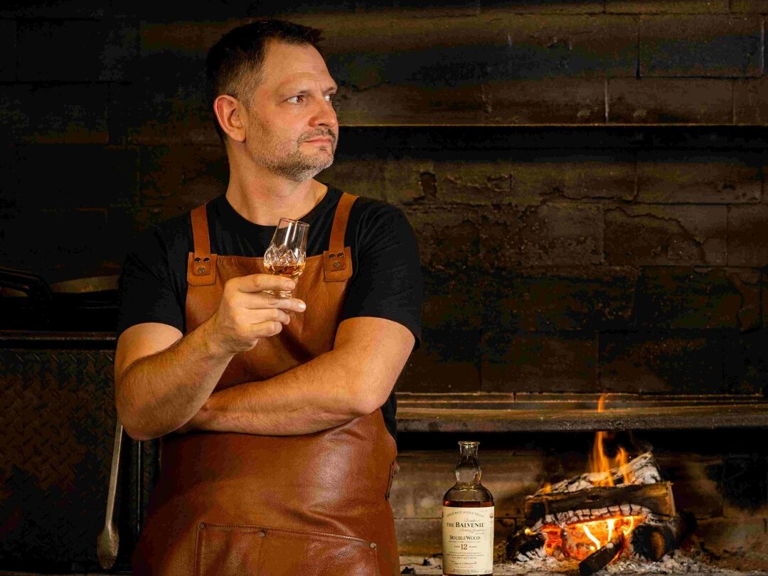 A man holds a glass of amber liquid, arms crossed, wearing a brown apron; he stands before a burning fireplace in a rustic brick setting