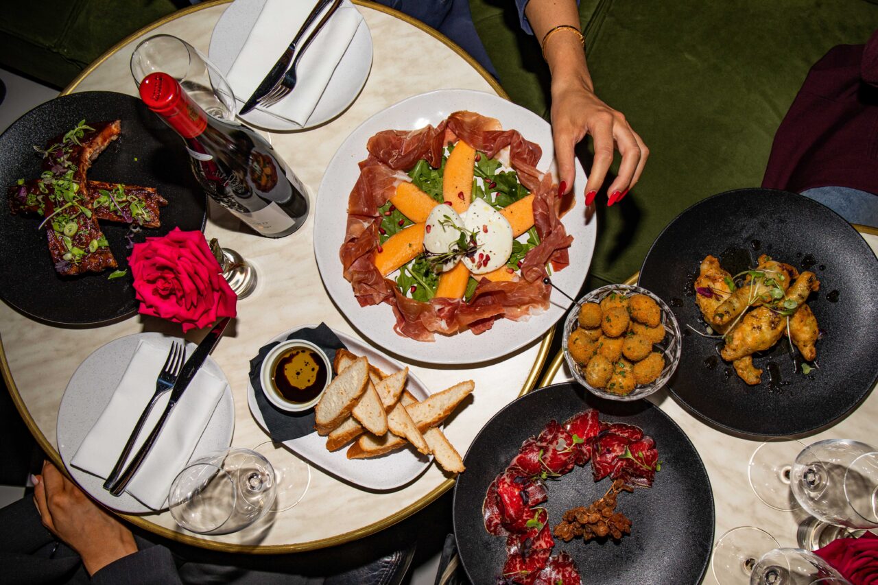 A prosciutto-melon-mozzarella salad is arranged on a plate; a hand reaches for it; the setting is a table with other dishes and wine glasses