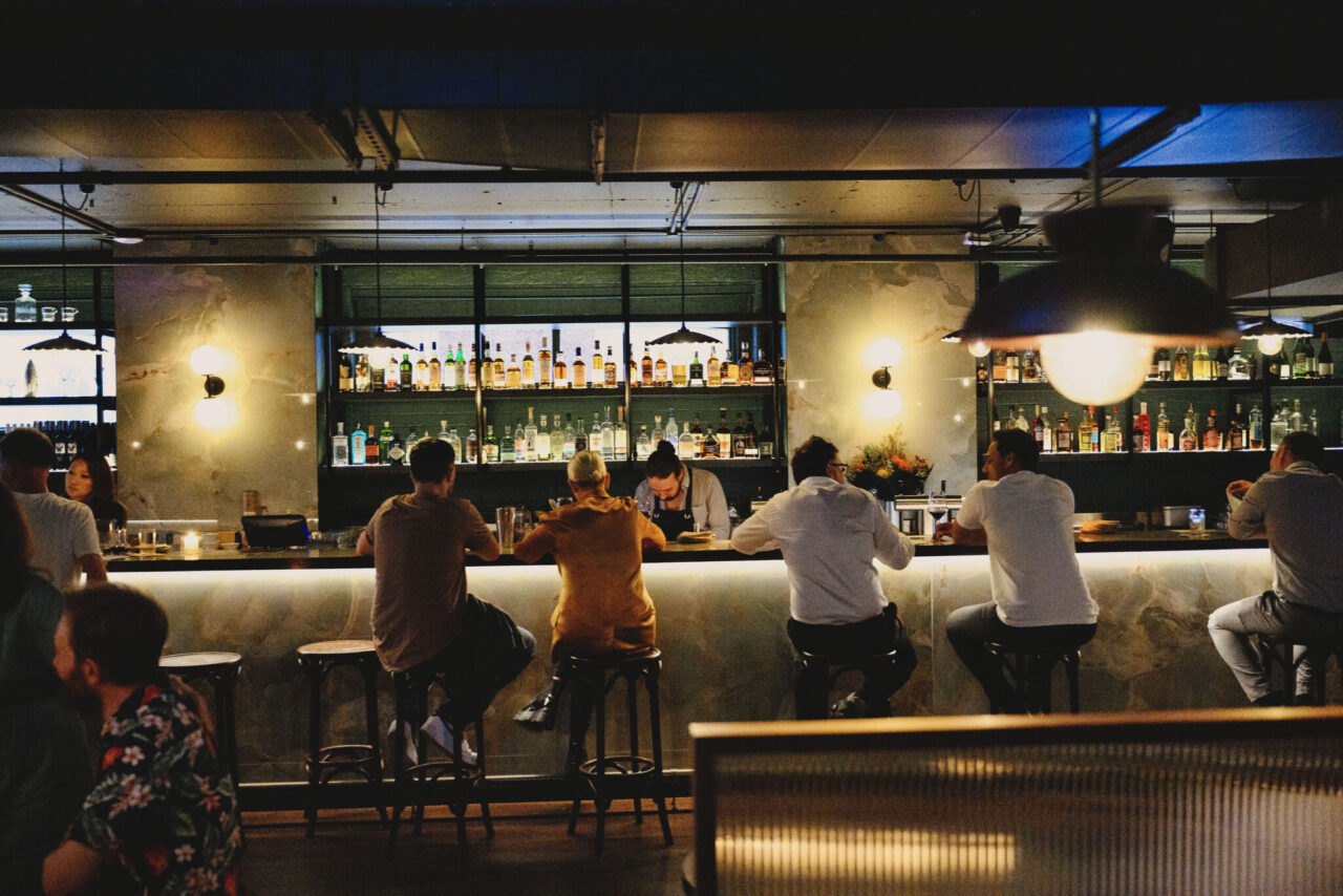 Patrons sit at a bar, facing a bartender, in a dimly lit, upscale establishment