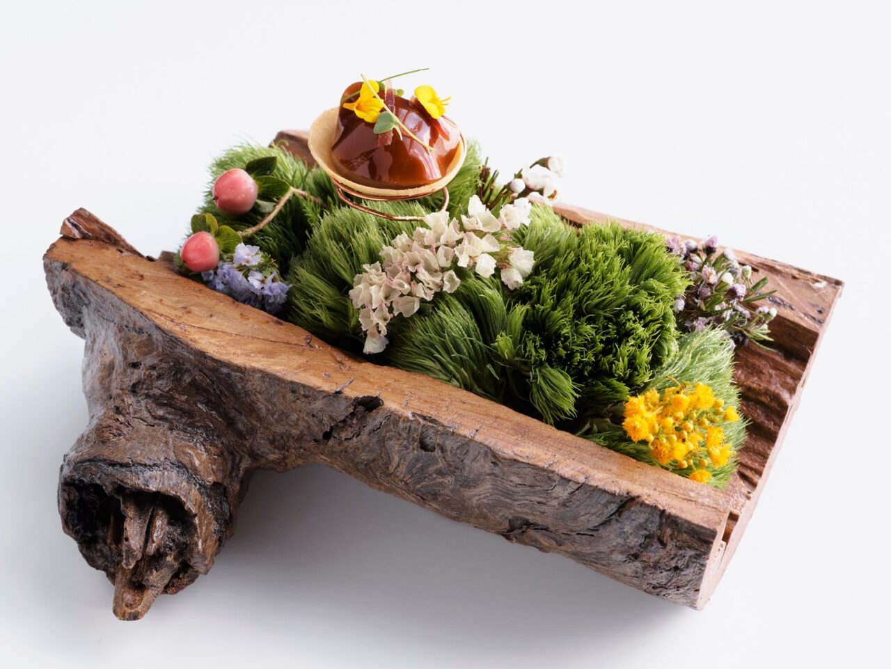 A caramel-colored dessert sits atop a small pastry, nestled within a bed of greenery in a rustic wooden trough