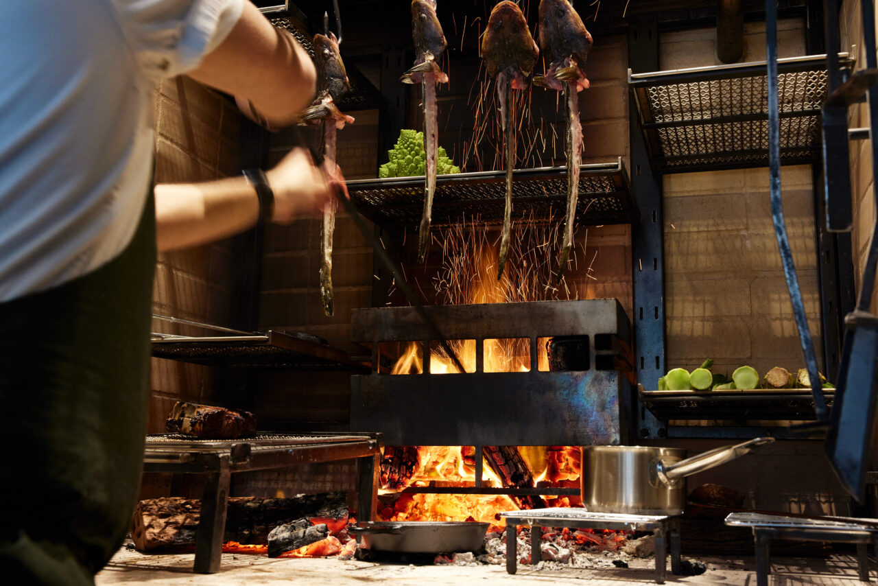 A chef manipulates fish on skewers over an open fire in a restaurant kitchen