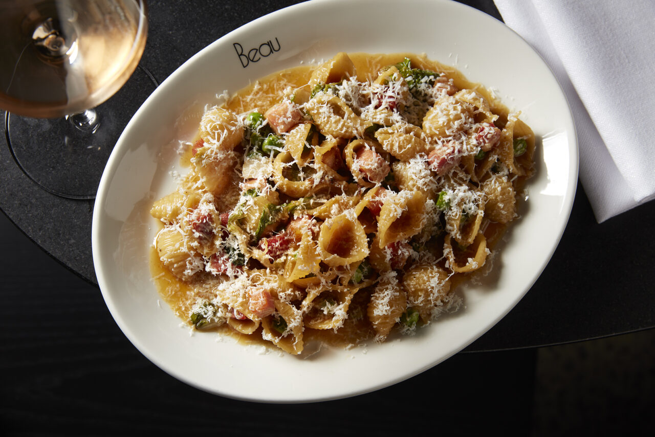 Shell pasta, topped with cheese and peas, is served on a white plate; a glass of rosé sits nearby on a dark table