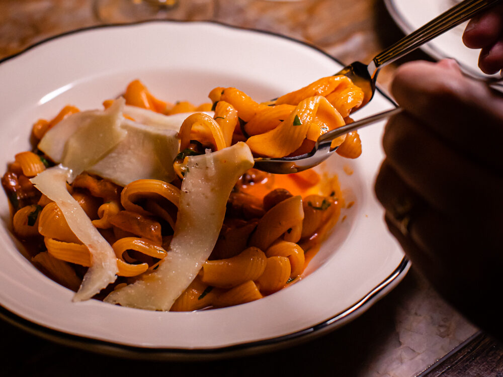 Orange pasta, topped with parmesan, is being served with tongs from a white bowl