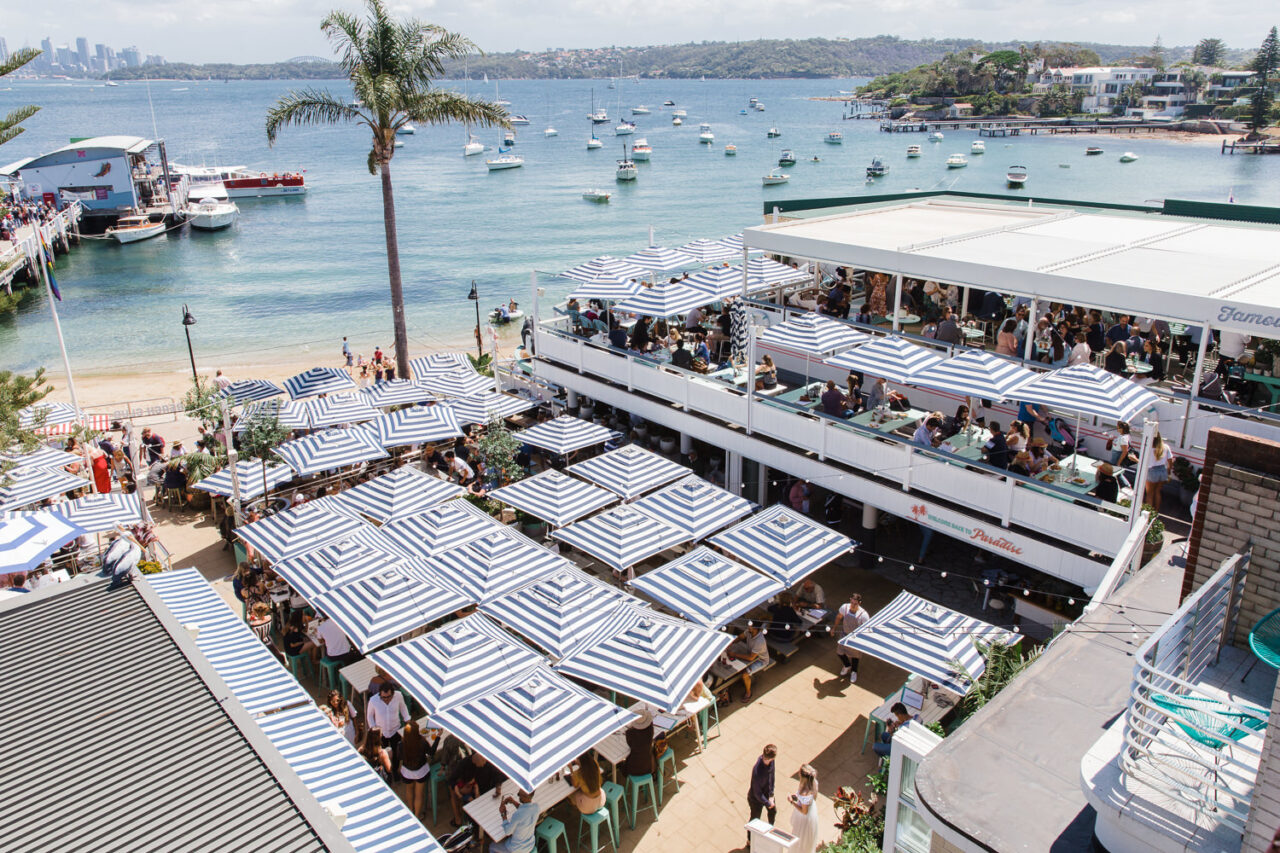 Numerous blue-and-white striped umbrellas shade patrons seated at tables in an outdoor beachfront restaurant