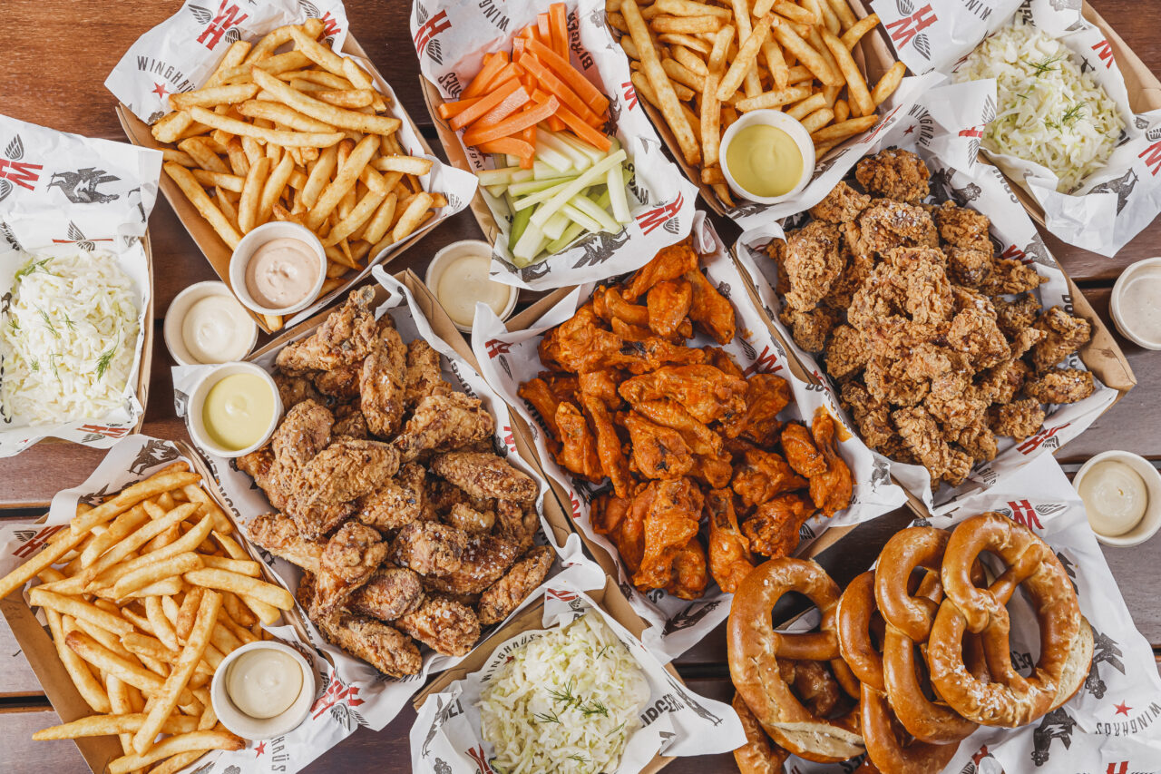 Several containers of fried chicken wings, french fries, and pretzels; plus sides of coleslaw and raw vegetables, are arranged on a wooden surface