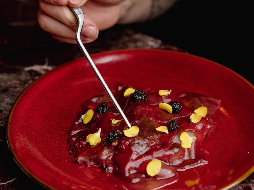 A pair of tweezers places small yellow petals onto a dark red beet carpaccio garnished with black caviar, served on a red plate
