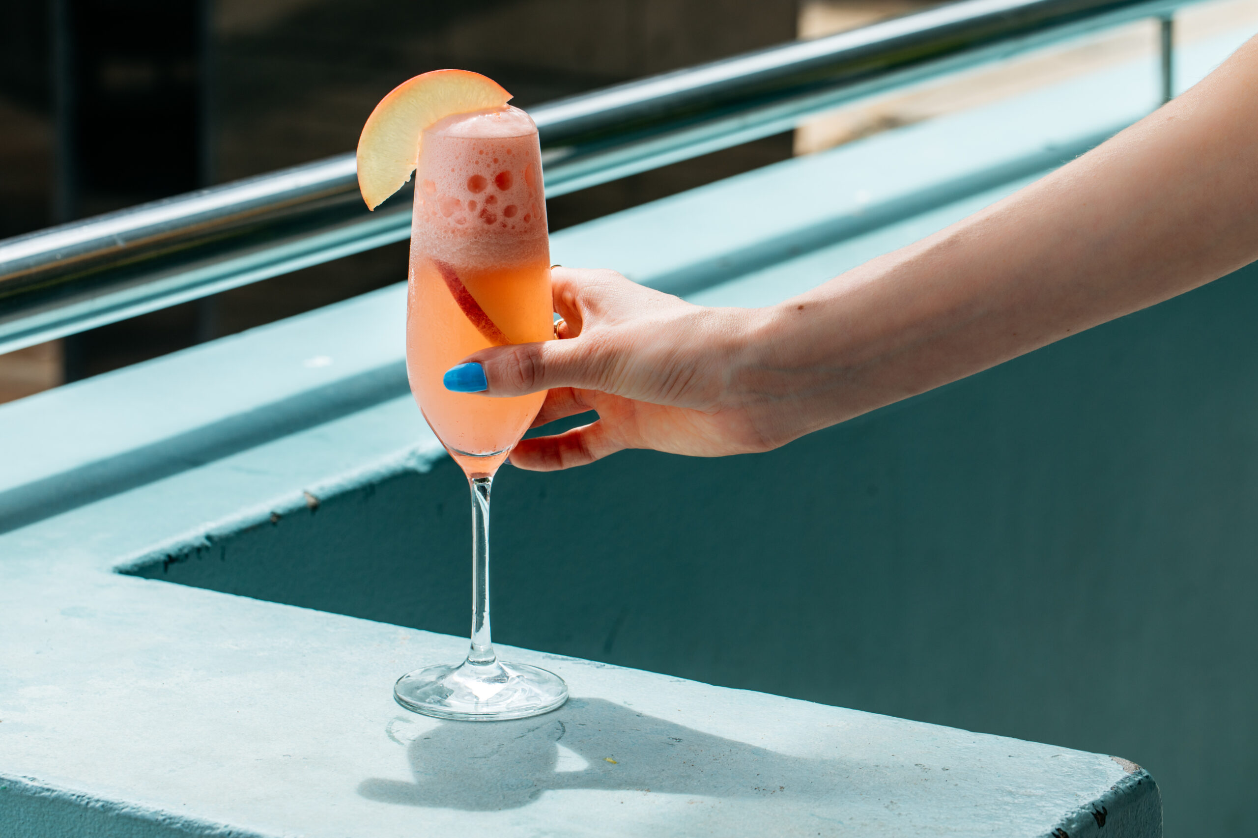 A peach-colored bubbly beverage, garnished with a peach slice, is held in a champagne flute by a hand