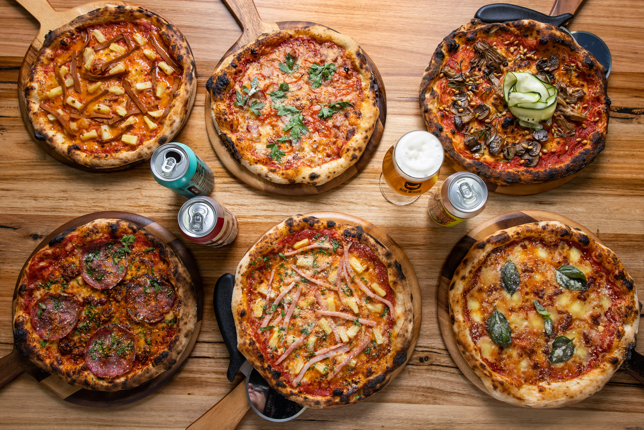 Six pizzas, arranged on wooden boards, are displayed on a wood table alongside beer and canned drinks