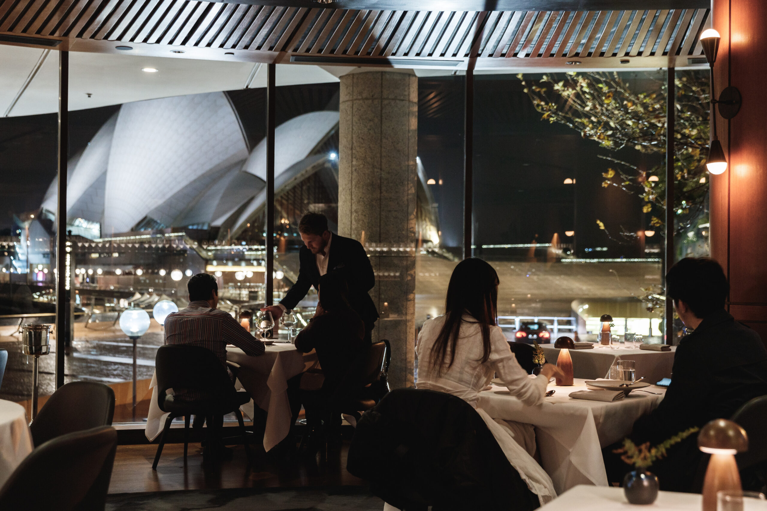 Diners are seated at tables, a waiter serves, overlooking a nighttime cityscape featuring the Sydney Opera House