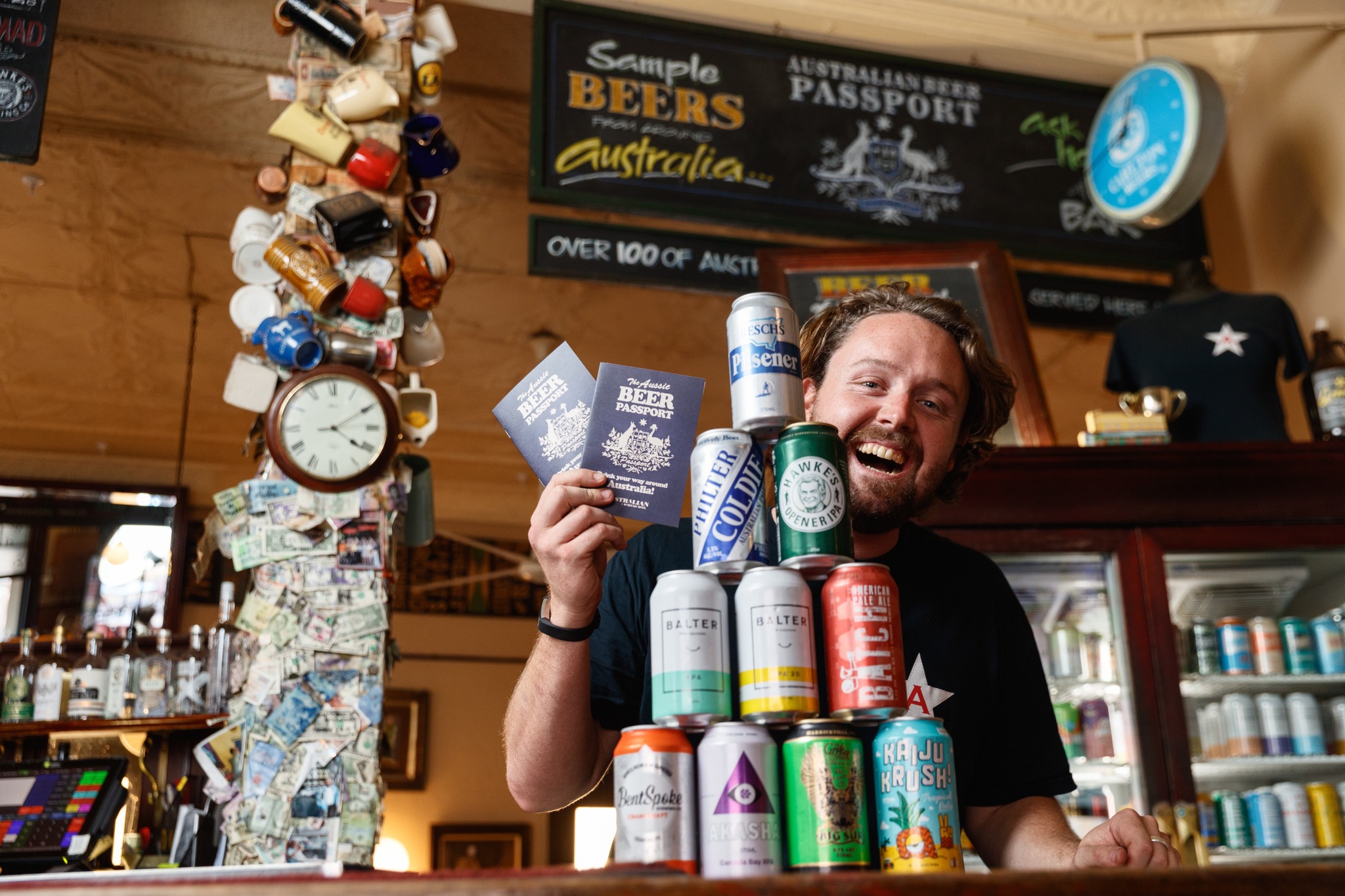 A man displays two "Aussie Beer Passport" booklets and a stack of various beer cans in a bar setting