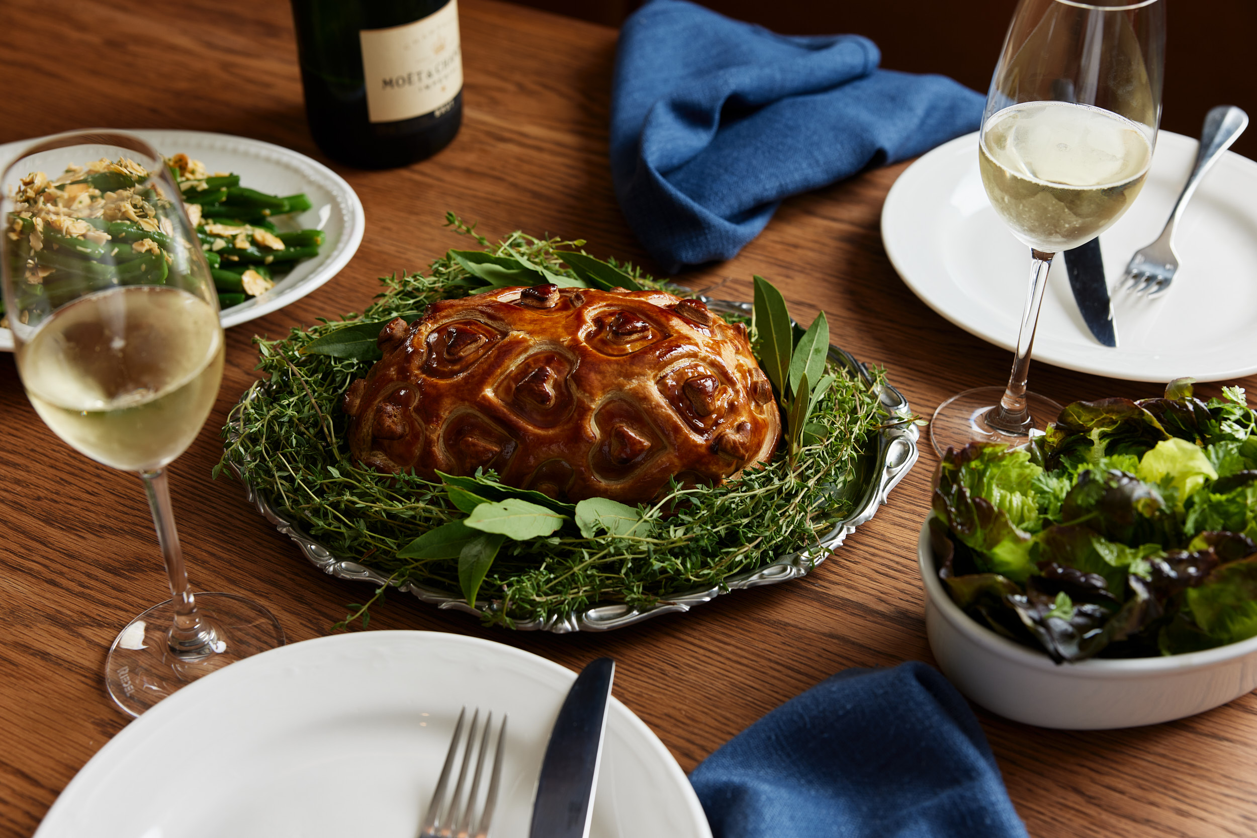A large, golden-brown meat pie rests on a silver platter, surrounded by herbs