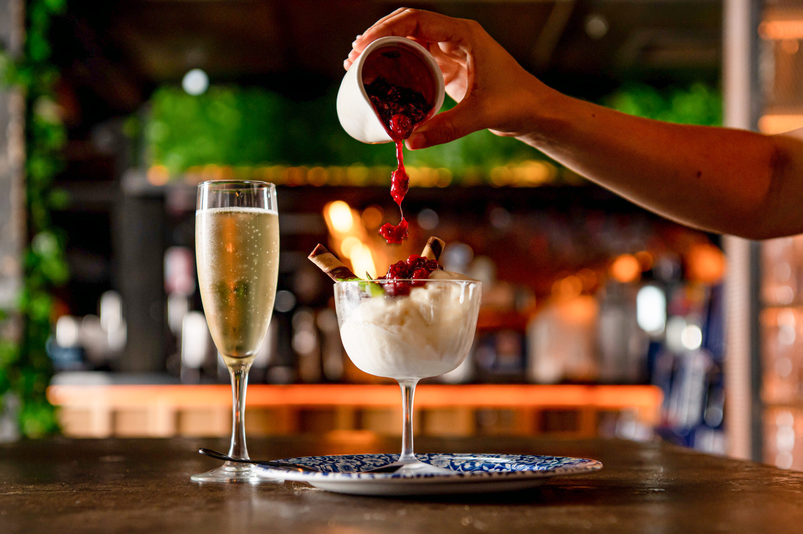 A hand pours a red berry sauce onto vanilla ice cream with a wafer, in a stemmed glass, on a bar top, next to a champagne flute