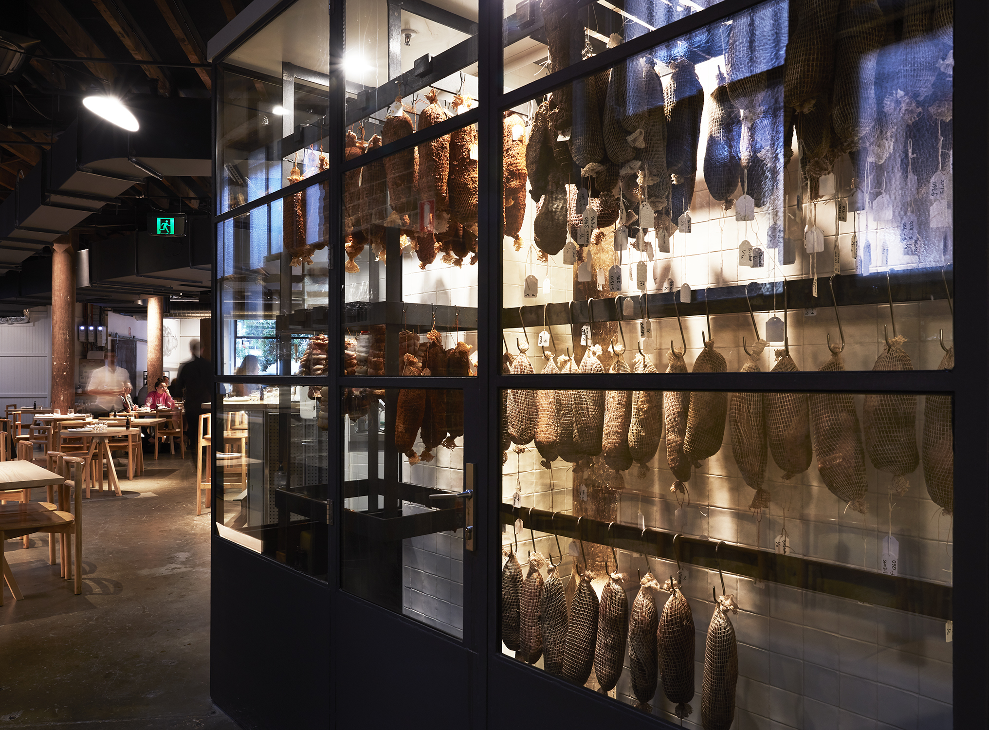 Cured meats hang in mesh bags from hooks inside a glass-fronted display case within a restaurant