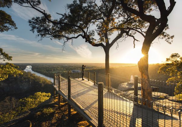 A person photographs a sunlit river valley from an elevated wooden platform; a large tree shades part of the platform