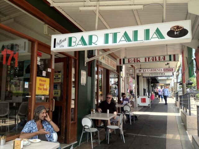 A man sits outside Bar Italia, a cafe, on a sunny day, talking on his phone