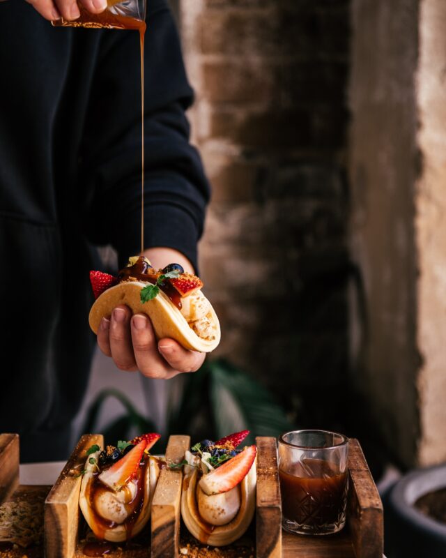 A person pours sauce over a pancake taco topped with fruit; two similar tacos sit on a wooden stand nearby