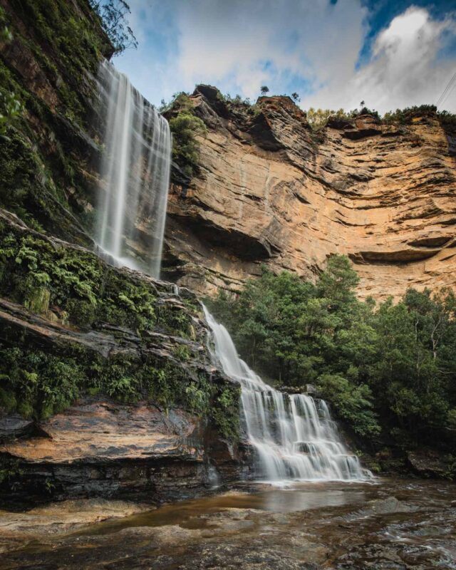 A waterfall cascades down a steep, layered sandstone cliff face, surrounded by lush green vegetation in a mountainous landscape