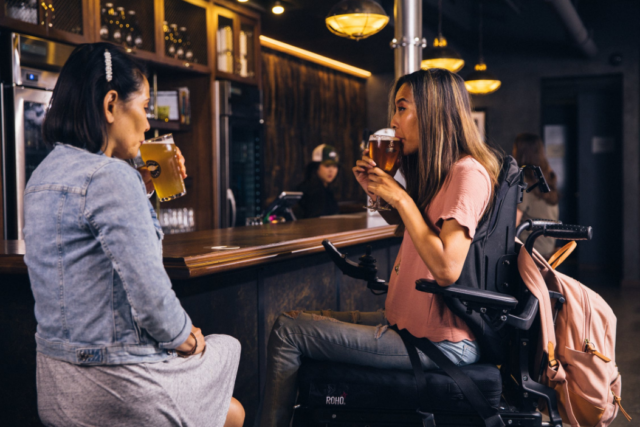 Two women sit at a bar, drinking beer