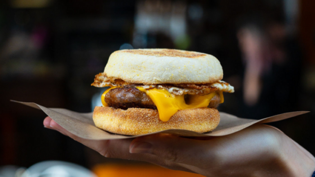 A breakfast sandwich, being held, consists of a sausage patty, fried egg, and melted cheese between English muffins