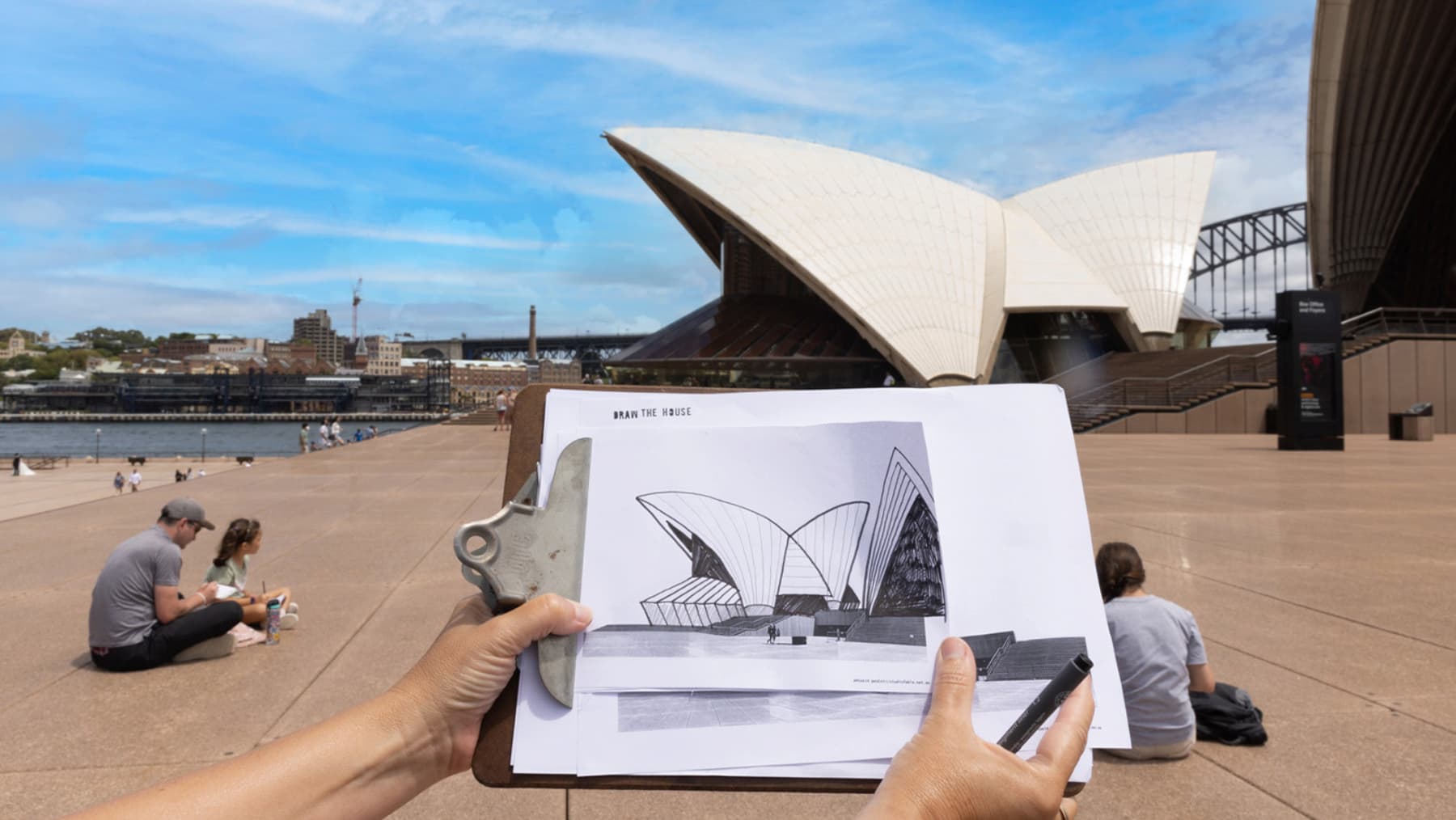 A clipboard displays a sketch of the Sydney Opera House; a person holds it, drawing in front of the actual building