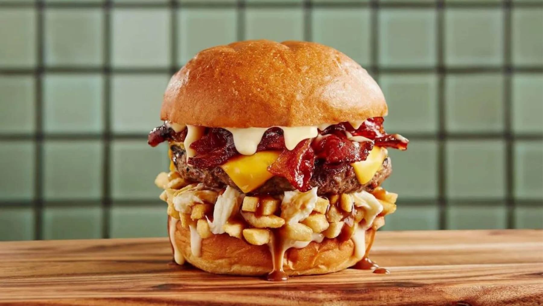 A large burger, topped with bacon, cheese, fries, and sauce, sits on a wooden board against a tiled background