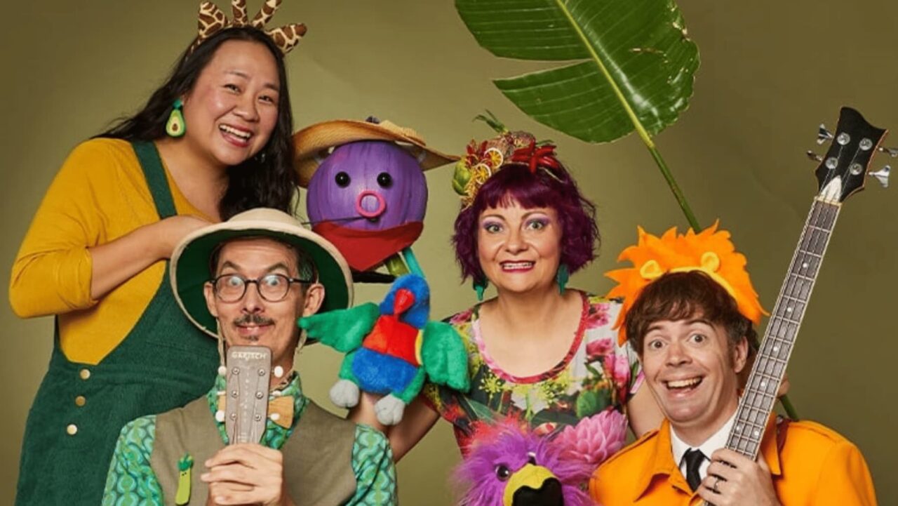 Four adults, wearing whimsical costumes and holding puppets and instruments, pose together against a muted green backdrop