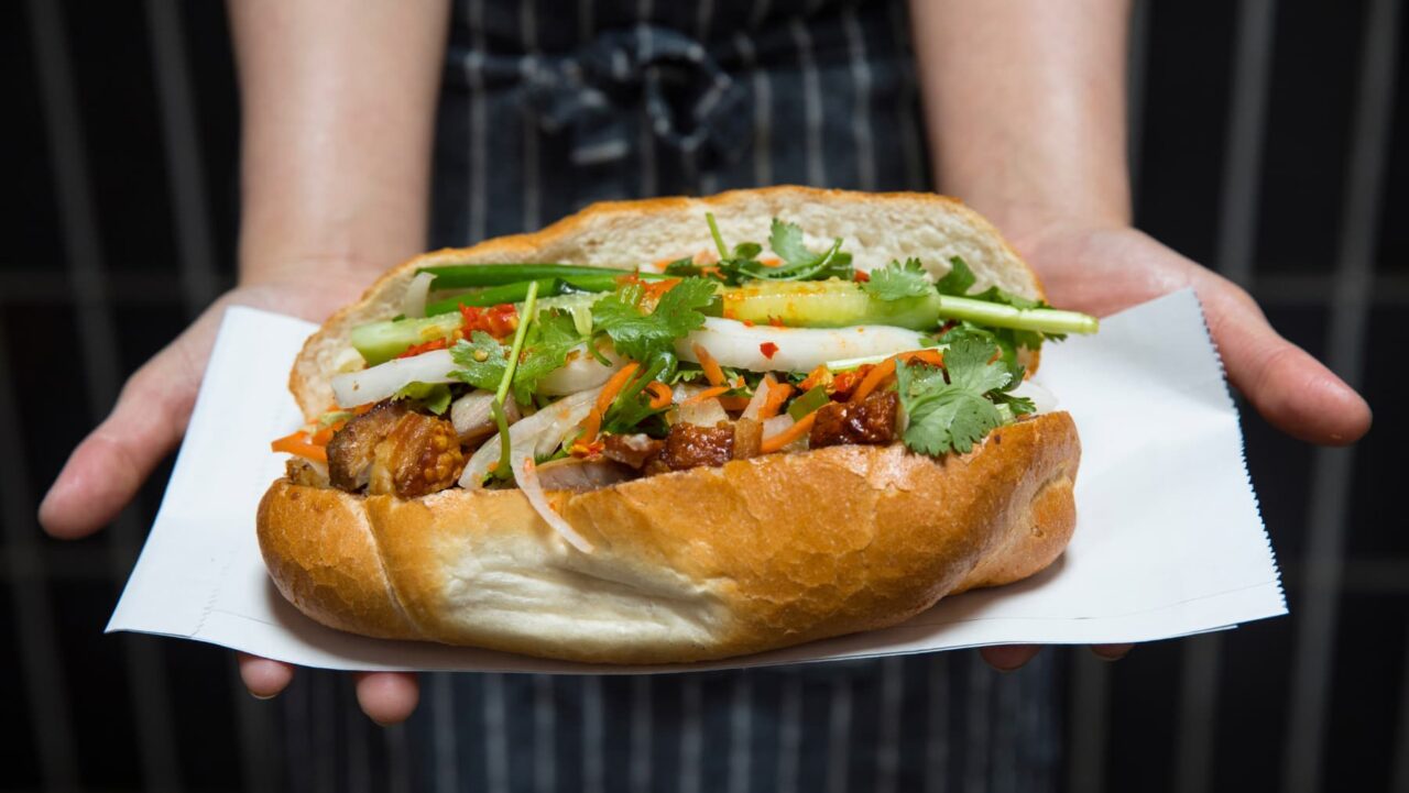 A banh mi, filled with meat and fresh vegetables, is being presented on a white paper liner by a person's hands