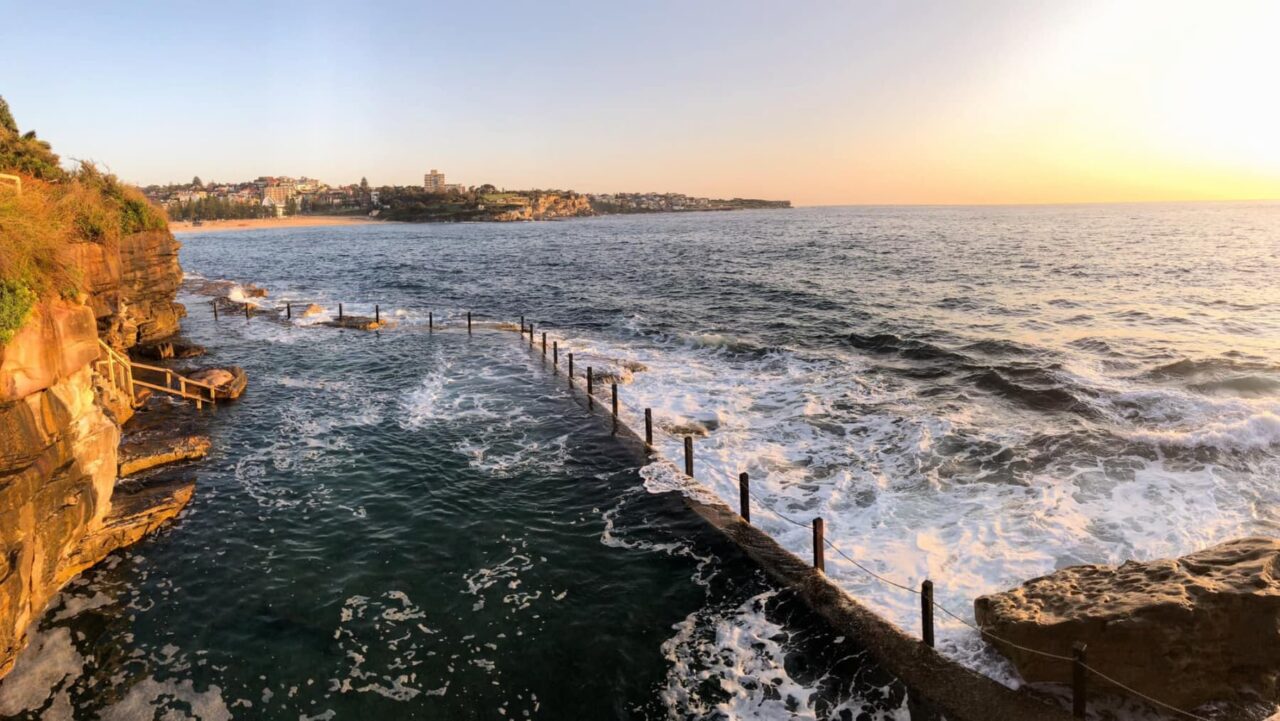 An ocean pool is partially filled with water, bordered by a low rock wall and wooden posts; waves crash against the wall at sunset, with a town visible in the distance