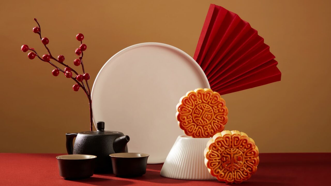 Two mooncakes rest on a small, white stand; a teapot and teacups sit beside a large white plate and a red fan, all arranged on a red surface
