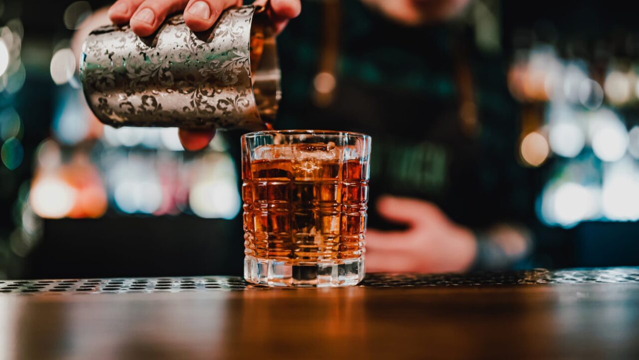 A bartender pours amber liquid from an ornate metal shaker into a rocks glass filled with ice at a bar