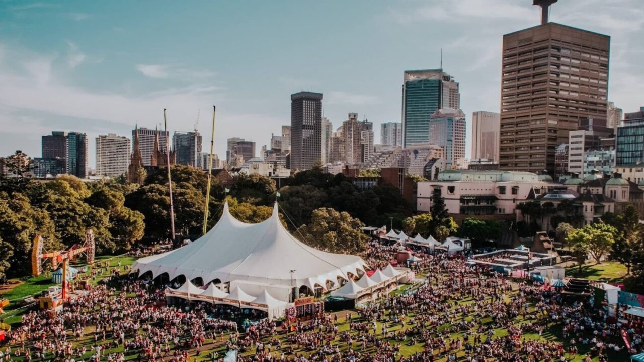 A large white tent sits amidst a crowd in a park, with a city skyline as a backdrop